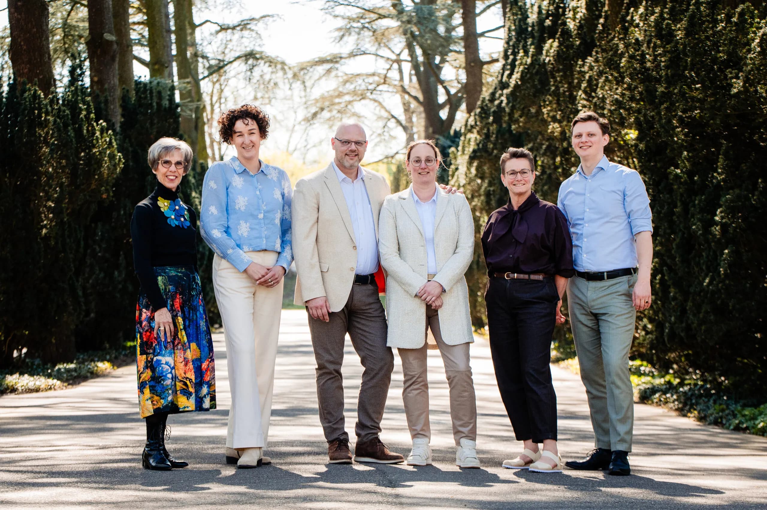 Het team van Els de Zanger Uitvaartbegeleiding, zes personen naast elkaar op een pad tussen hoge bomen