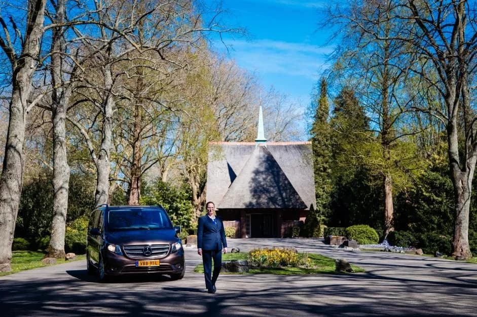Een uitvaartauto op het pad voor een rietgedekte kapel, met een uitvaartbegeleider die ernaartoe loopt