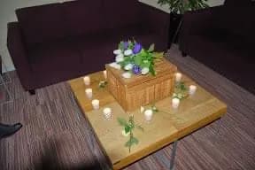 Een klein licht kistje op een lage tafel in de familiekamer, omringd door brandende kaarsjes en bloemen