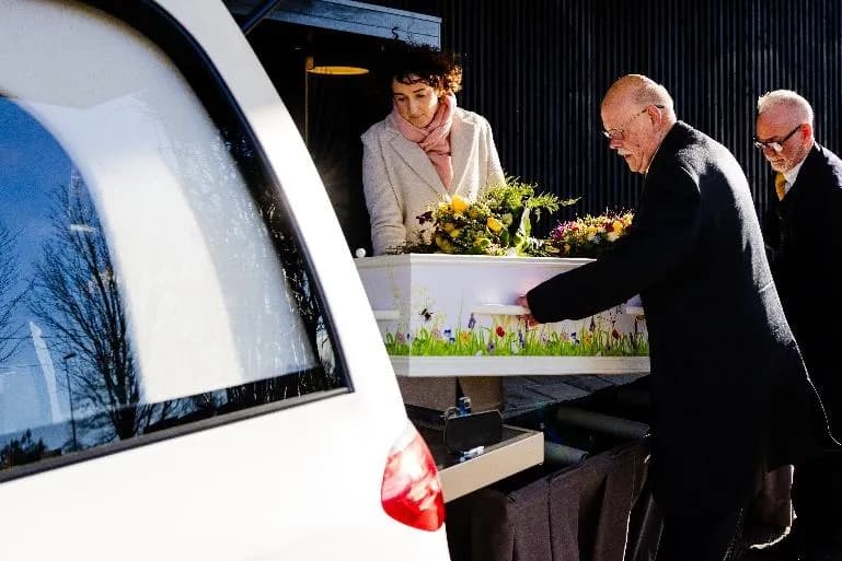 Annemarie Voogt en twee dragers schuiven een witte kist met bloemen in de rouwauto